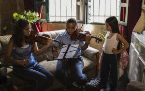  Dissandra and friend rehearse violin, sitting while Dissandra's sister watches them