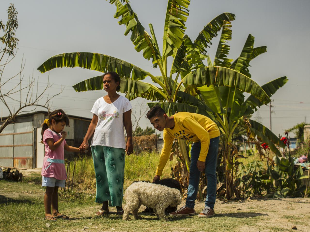 Production Still from Children of Las Brisas. Two adults and a child stand behind a tree in Venezuela. There is also a small dog in the photo.